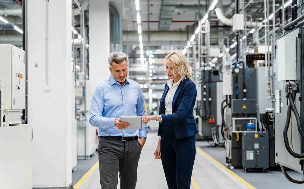 Ein Mann und eine Frau in Businesskleidung besprechen etwas auf einem Tablet in einer Produktionshalle.