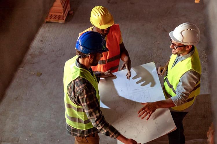 Drei Männer in Bauschutzkleidung besprechen einen Plan auf einer Baustelle.