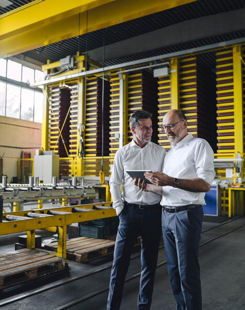 Zwei Männer in weißen Hemden unterhalten sich und gucken auf ein Tablet in einer Betriebswerkstatt.