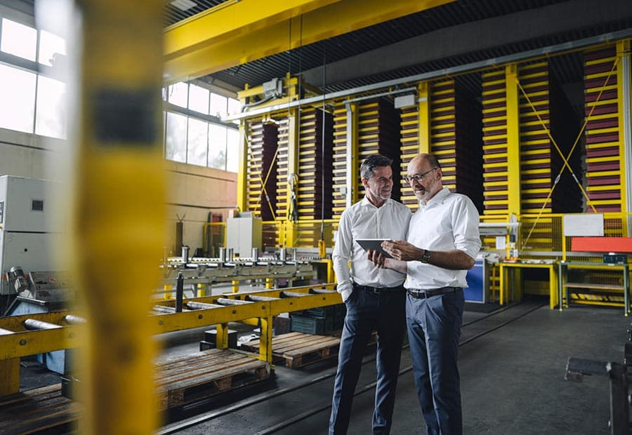 Zwei Männer in weißen Hemden unterhalten sich und gucken auf ein Tablet in einer Betriebswerkstatt.
