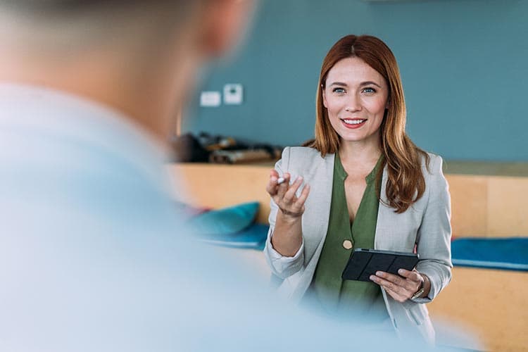 Eine Frau im Blazer erklärt einem Mann etwas und hält ein Tablet in der Hand.