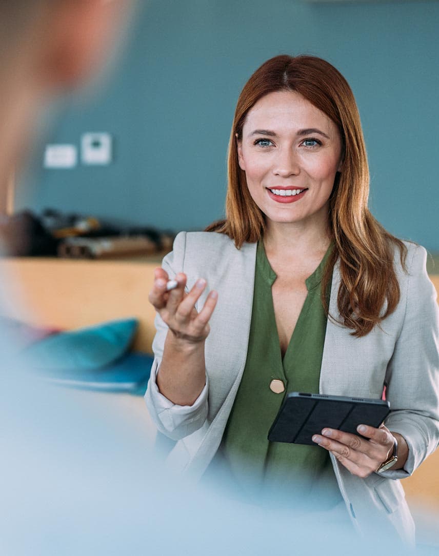 Eine Frau im Blazer erklärt einem Mann etwas und hält ein Tablet in der Hand.
