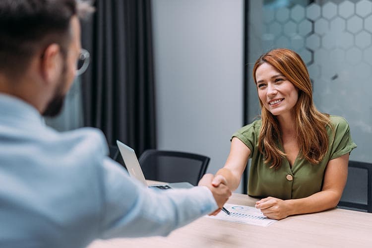 Eine Frau sitzt an einem Meetingtisch und reicht ihrem Kollegen die Hand.
