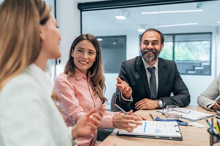 Ein Mann und eine Frau lachen gemeinsam mit einer Kollegin an einem Meetingtisch.