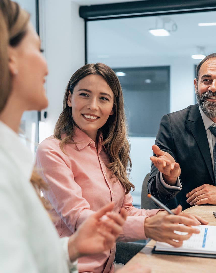 Ein Mann und eine Frau lachen gemeinsam mit einer Kollegin an einem Meetingtisch.