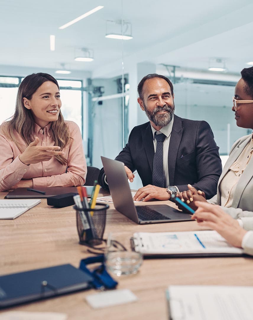 Ein Mann und eine Frau lachen gemeinsam mit KollegInnen an einem Meetingtisch, darauf steht ein Laptop.