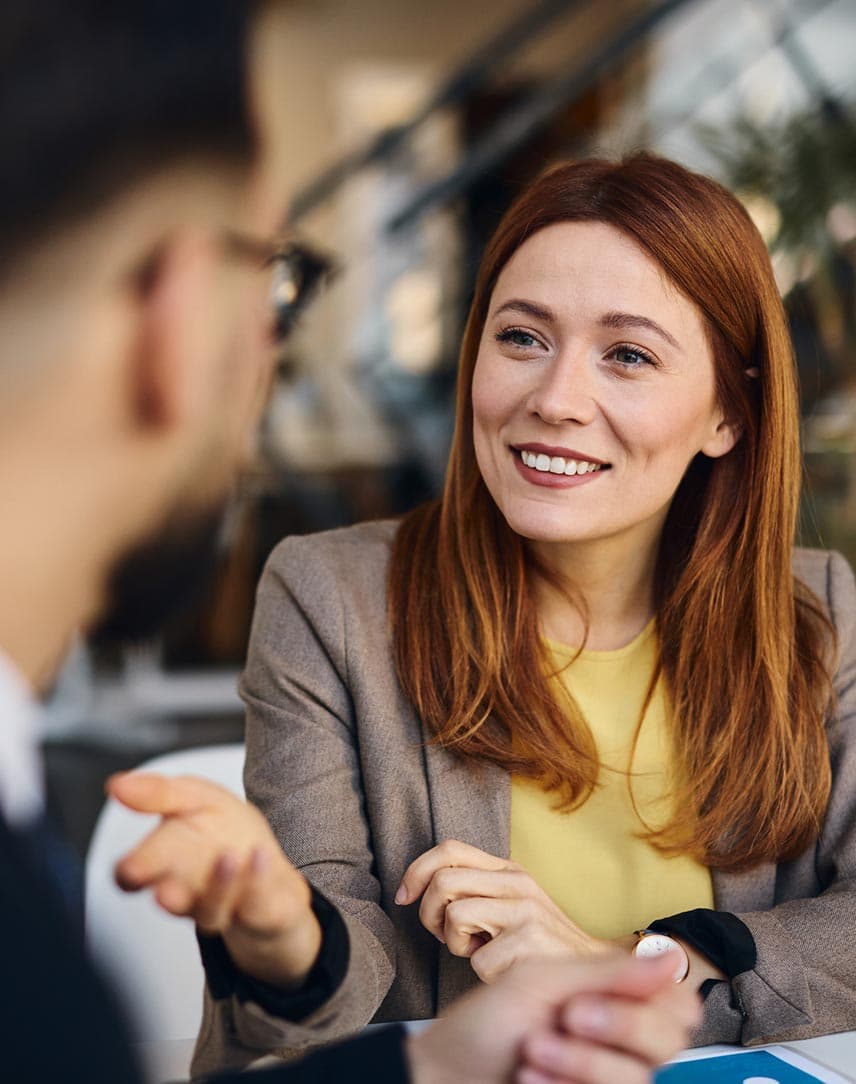Eine Frau in Businesskleidung sitzt an einem Bürotisch und unterhält sich mit einem Mann im Anzug.