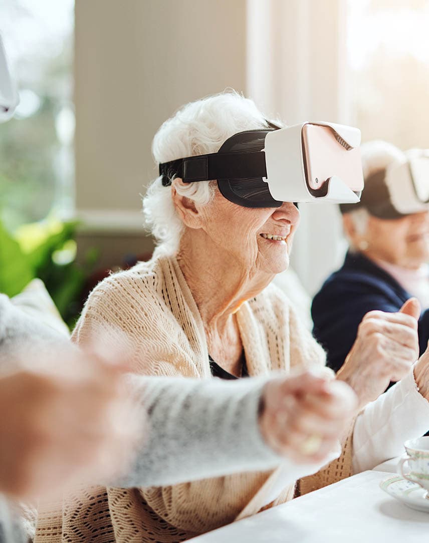 Drei Seniorinnen und Senioren sitzen an einem Tisch in einem Pflegeheim, haben Virtual Reality Brillen mit Smartphones auf und spielen gemeinsam Online-Spiele.