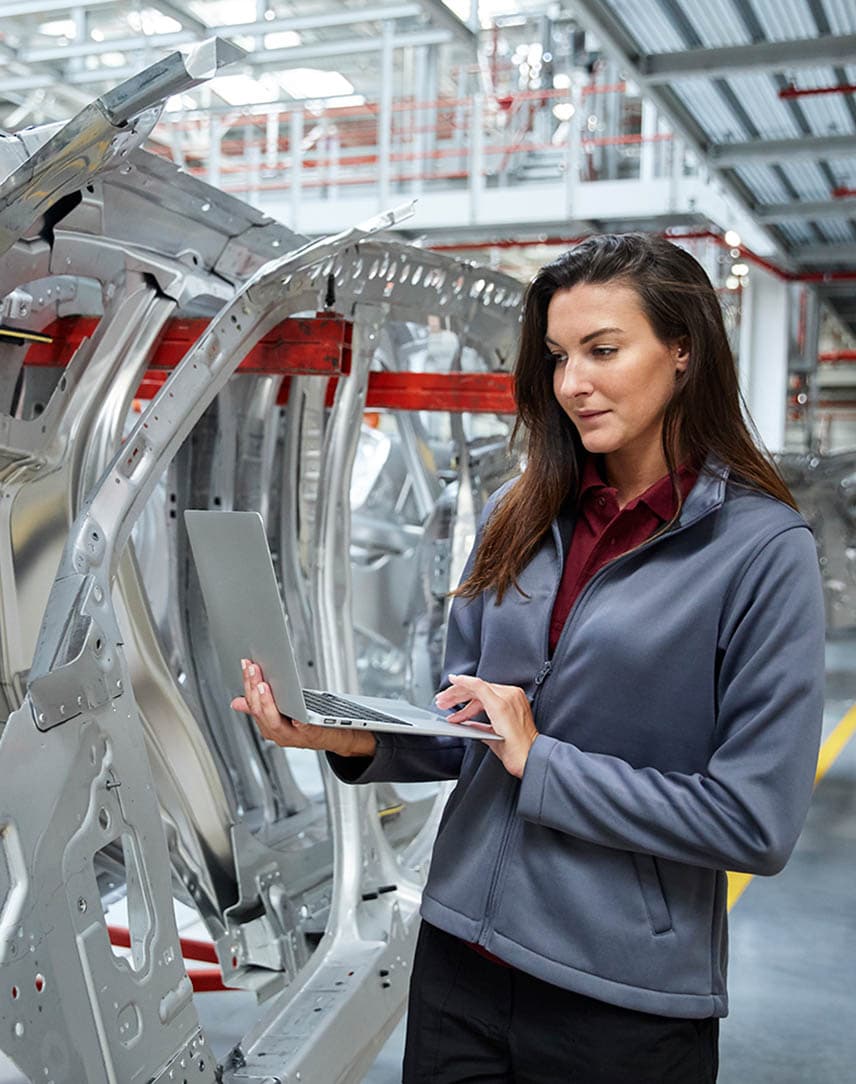 Eine Frau steht in einer Produktionshalle neben silbernen Rohlingen von Fahrzeugteilen. Sie hält in Laptop in der Hand und schaut darauf.