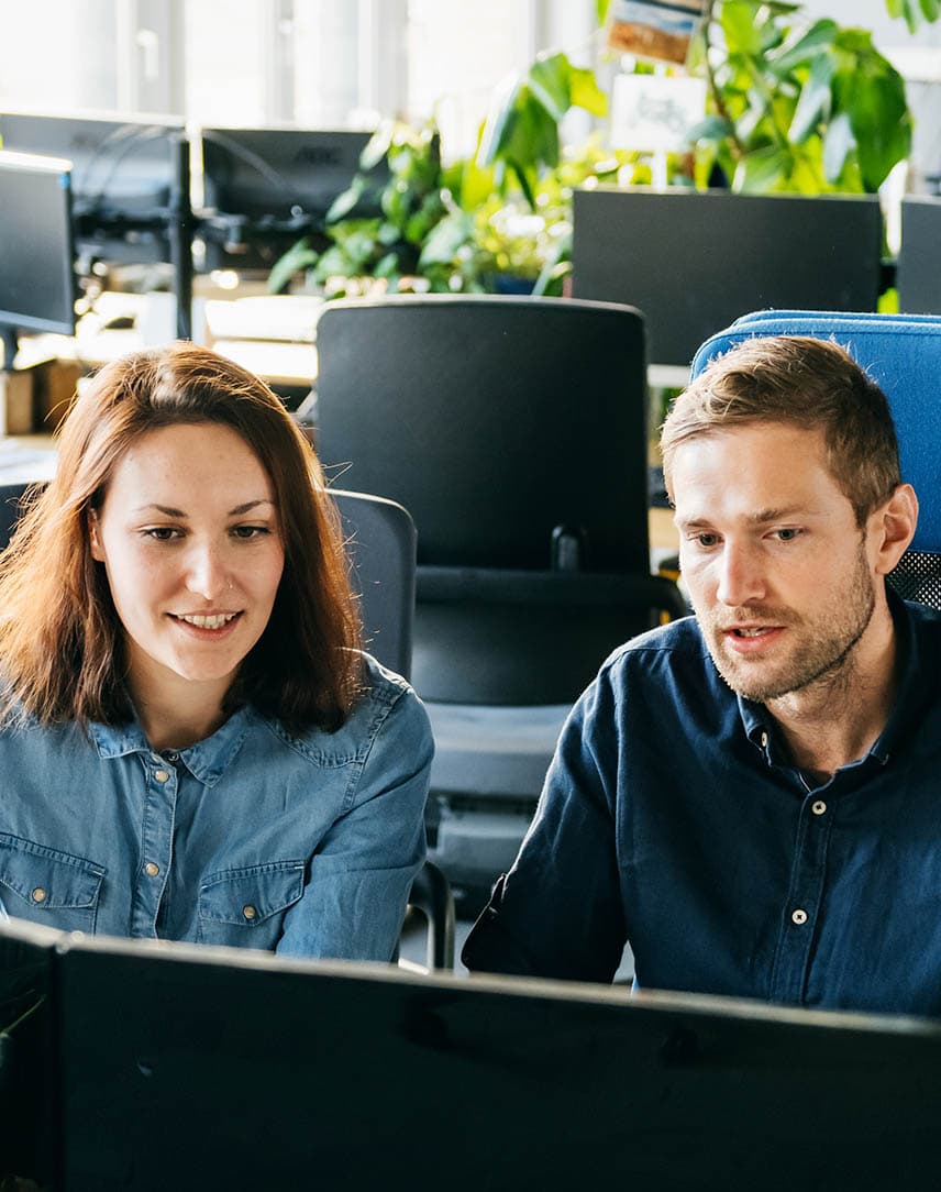 Ein Mann im dunkelblauen Hemd und eine Frau im hellblauen Hemd sitzen vor zwei Bildschirmen in einem Büro.