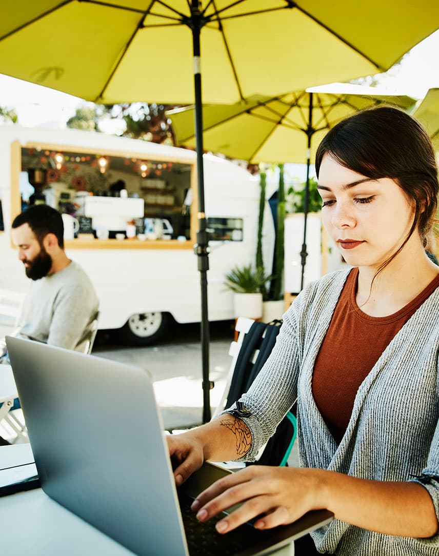 Eine junge Frau arbeitet auf einer sonnenbeschienen Cafe-Terrasse unter einem Sonnenschirm an einem Laptop, im Hintergrund ein Mann