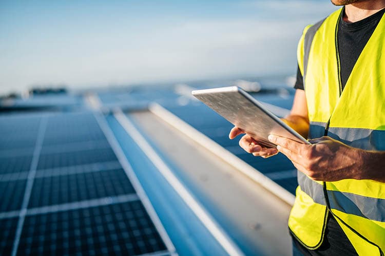 Ein Techniker mit Helm und Warnweste steht mit Tablet auf einem Dach, auf dem PV-Module installiert sind.
