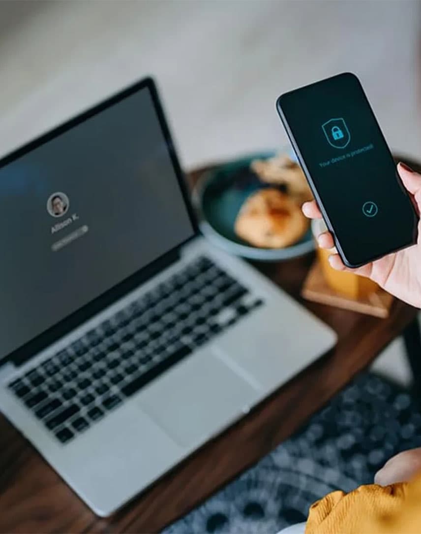 Eine Frau an einem Laptop und mit einem Handy in der Hand, auf dem ein Schloss als Sicherheitssymbol zu sehen ist