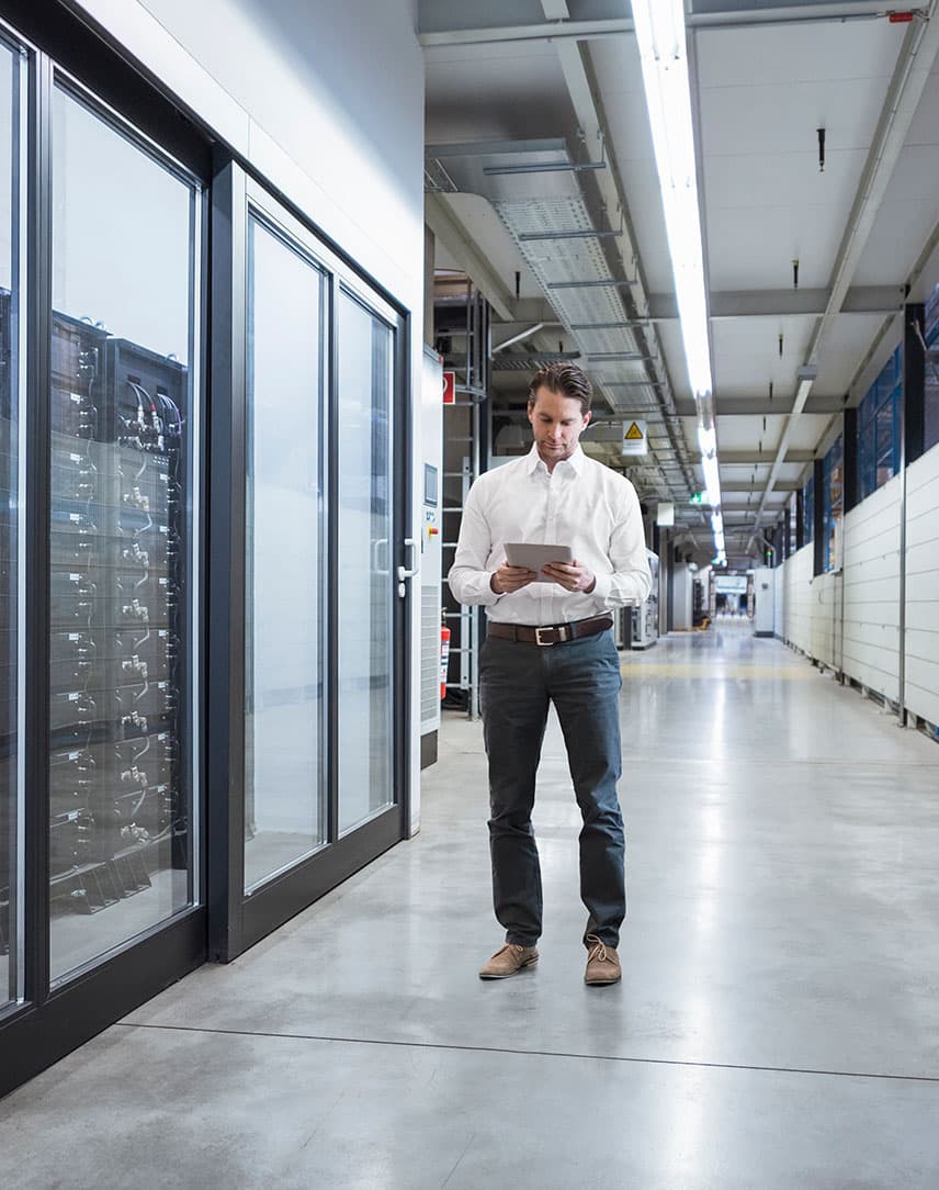 Ein Mann im weißen Hemd steht mit einem Tablet neben einem Serverbereich in einer Fabrik.