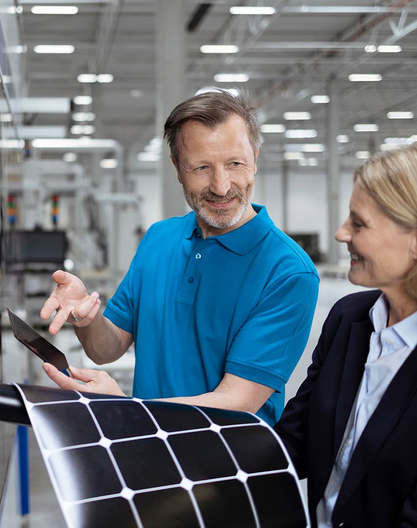 Ein Mann zeigt einer Frau in einem Fabrikgebäude ein Bauteil, die Frau hat eine dehnbare PV-Fläche in den Händen.