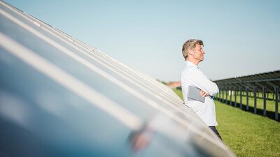Ein Mann im weißen Hemd mit Tablet lehnt an einer PV-Anlage auf grüner Wiese.