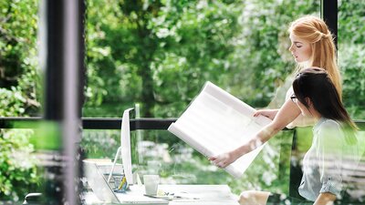 Webinar: Korridorphase CO2-Preis Zwei Kolleginnen bei der Büroarbeit vor einer Fensterfront, hinter der Bäume zu sehen sind.