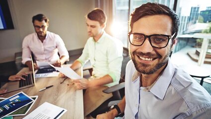 Ein Mann mit Brille an einem Bürotisch, im Hintergrund zwei Kollegen