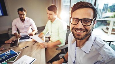 Ein Mann mit Brille an einem Bürotisch, im Hintergrund zwei Kollegen