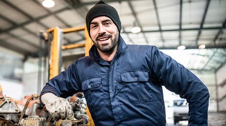Ein junger Mann im Blaumann lehnt an einem unmontiertem Motor in einer Werkshalle.