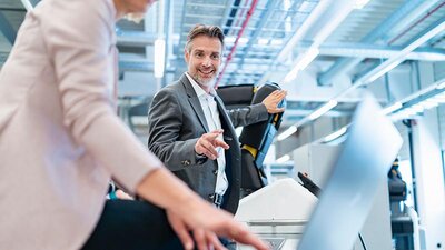 Ein lächelnder Mann im grauen Anzug deutet in einer Werkhalle auf einen Laptop, den eine Frau in Benutzung hat.
