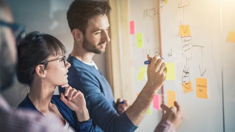 Mann, Frau und andere Personen beschriften Post-its an eine Whiteboard