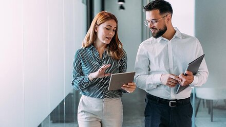 Eine Frau läuft neben einem Mann in einem Büro und erklärt ihm etwas anhand eines Tablets.