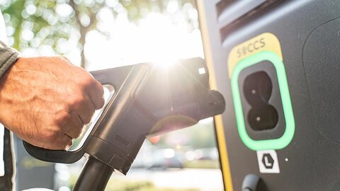 Ein Hand mit Stecker und eine Ladesäule, im Hintergrund eine sonnenbeschienene Straße