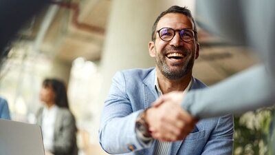 Ein Mann mit Brille und Sakko begrüßt jemanden per Handschlag und freut sich dabei.