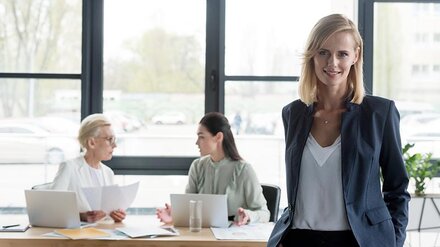 Eine Businessfrau in einem Büro, im Hintergrund zwei weitere Mitarbeiterinnen an einem Bürotisch