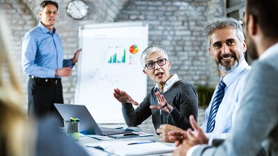 Geschäftsleute diskutieren untereinander. Im intergrund steht ein Seminarleiter an einem Flipchart