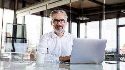 Eine Mann im weißen Businesshemd sitzt vor einem Laptop und lächelt.