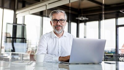 Eine Mann im weißen Businesshemd sitzt vor einem Laptop und lächelt.