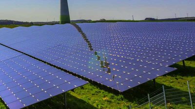 Eine PV-Anlage auf einer grünen Wiese, im Hintergrund eine Windenergieanlage
