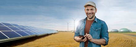 Direktvermarktung mit EWE business Ein junger Landwirt auf einem Feld, links eine PV-Anlage im Hintergrund Windräder und eine Biogasanlage