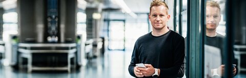 EWE business Terminmarktportal Ein Mann mit Handy, der in einem Büro an einer Glaswand lehnt