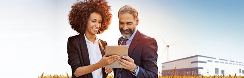 EWE business Flexload Eine Frau und ein Mann stehen in einem Getreidefeld und gucken auf ein Tablet, im Hintergrund ein Unternehmensgebäude und ein Windrad