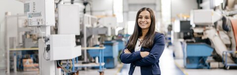 EWE business für Industrieunternehmen Eine Frau im blauen Blazer steht mit verschränkten Armen in einer Produktionshalle und lächelt.
