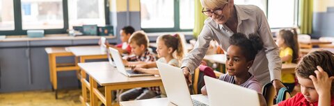 Chancen durch digitale Infrastruktur Eine Lehrerin erklärt Schülerinnen und Schülern etwas an Computern im Klassenraum.