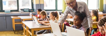 Chancen durch digitale Infrastruktur Eine Lehrerin erklärt Schülerinnen und Schülern etwas an Computern im Klassenraum.