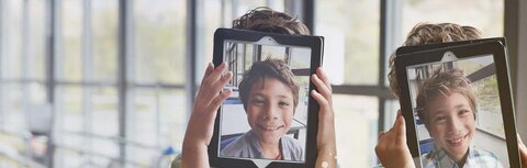 Geräte-Wirrwarr an der Schule Kindergesichter auf Tablets, die die Kinder hochhalten
