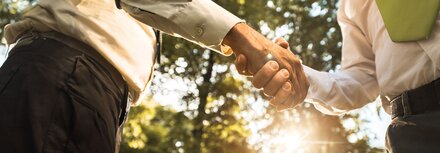 EWE business Eventreihe "Grüne Energie" Zwei Männer schütteln sich vertrauenvoll die Hände. Im Hintergrund eine natürliche Landschaft mit einer Sonne, die durch Baumkronen scheint.