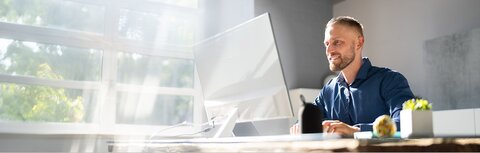 EWE business Events Ein Mann an einem Schreibtisch, der auf einen Bildschirm guckt, im Hintergrund eine helle Fensterfront
