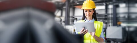 Asset Tracking für Logistik und Produktion Eine Frau mit Warnweste und Helm arbeitet mit dem Tablet in einer Industriehalle.