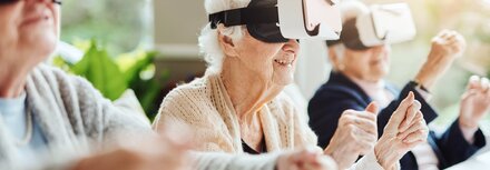 Digitalisierung in der Pflege Drei Seniorinnen und Senioren sitzen an einem Tisch in einem Pflegeheim, haben Virtual Reality Brillen mit Smartphones auf und spielen gemeinsam Online-Spiele.