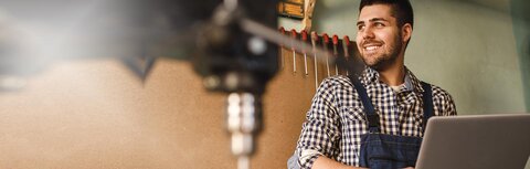 Online-Marketing für kleine Unternehmen Ein Handwerker hält ein Notebook in seiner Werkstatt und lächelt.