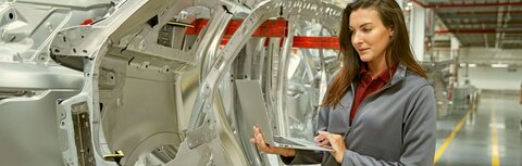 Standortvernetzung mit EWE Eine Frau steht in einer Produktionshalle neben silbernen Rohlingen von Fahrzeugteilen. Sie hält in Laptop in der Hand und schaut darauf.
