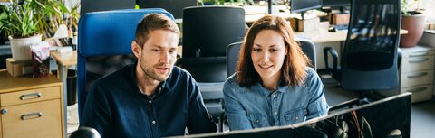 Teams-Telefonie für einen effizienten digitalen Austausch im Unternehmen Ein Mann im dunkelblauen Hemd und eine Frau im hellblauen Hemd sitzen vor zwei Bildschirmen in einem Büro.