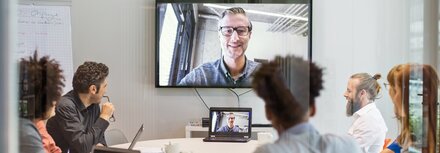 Videokonferenzen sicher machen In einem Besprechungsraum sitzen Frauen und Männer und nehmen an einer Videokonferenz mit einem Kollegen teil.
