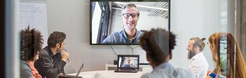 Videokonferenzen sicher machen In einem Besprechungsraum sitzen Frauen und Männer und nehmen an einer Videokonferenz mit einem Kollegen teil.